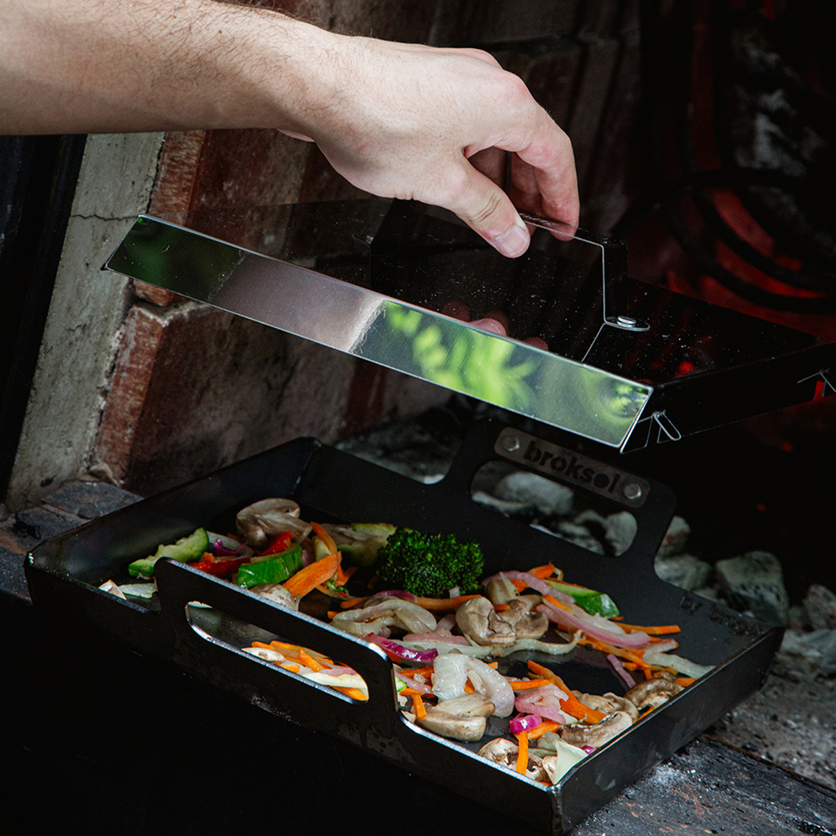 Mano levantando la tapa universal de una plancha con verduras salteadas