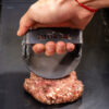 Mano aplastando carne picada con el smasher Brosen sobre la plancha