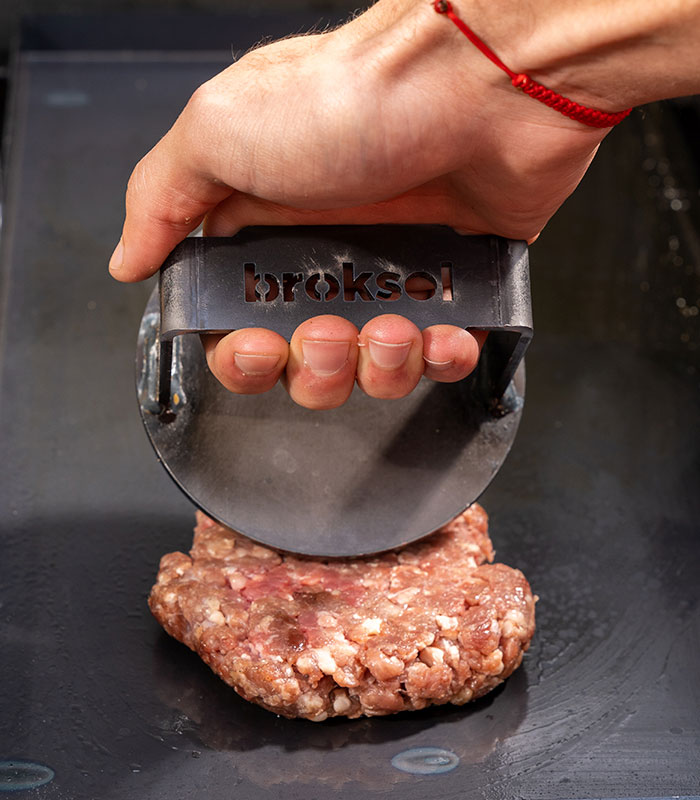 Mano aplastando carne picada con el smasher Brosen sobre la plancha